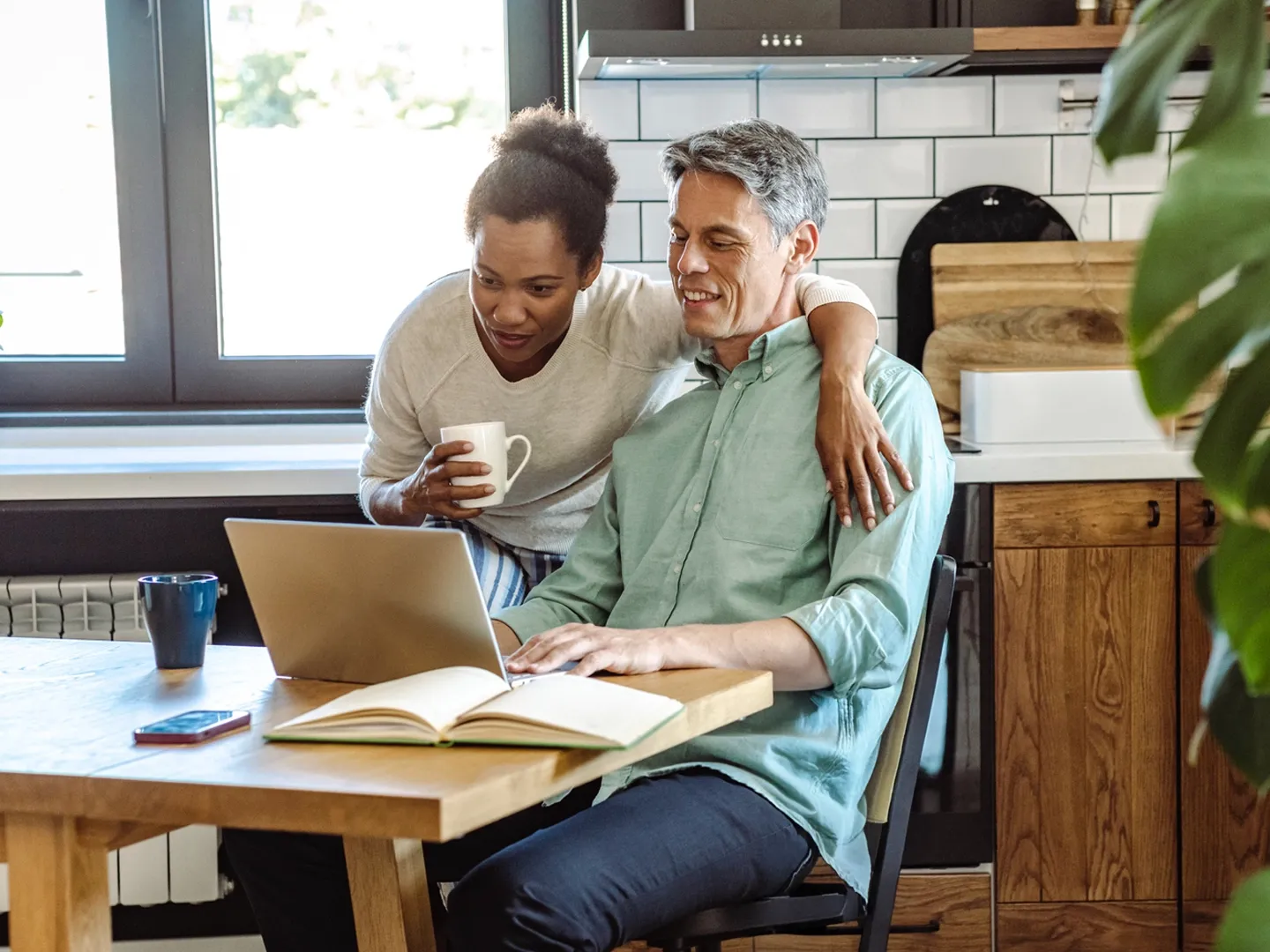A smiling man and woman look at a laptop screen together at a kitchen table.
